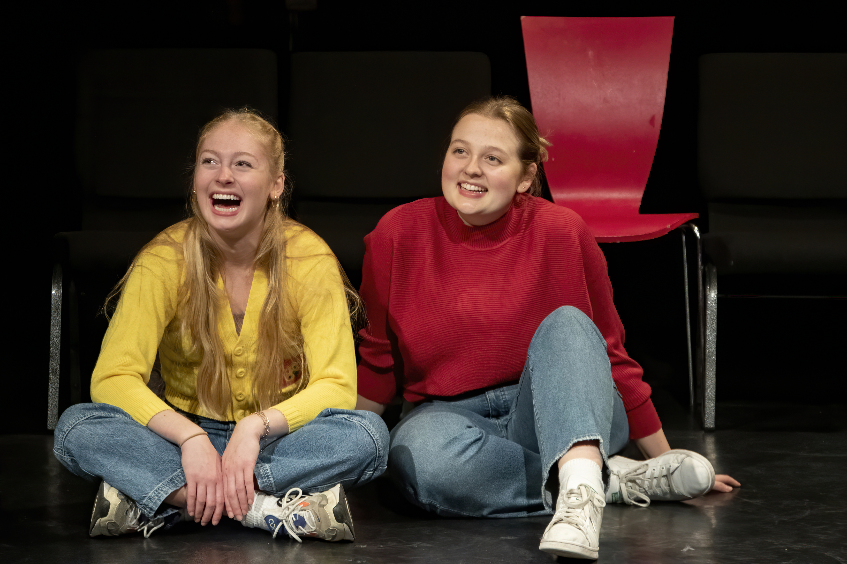 Two students seated on the floor of the black box theatre, smiling and laughing